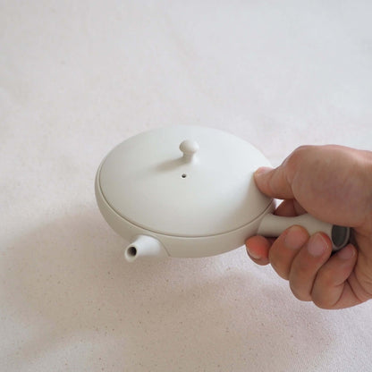 A Tokoname ware white flat teapot is held in the hand and tilted—its soft white appearance is a result of being shaped on a potter's wheel.