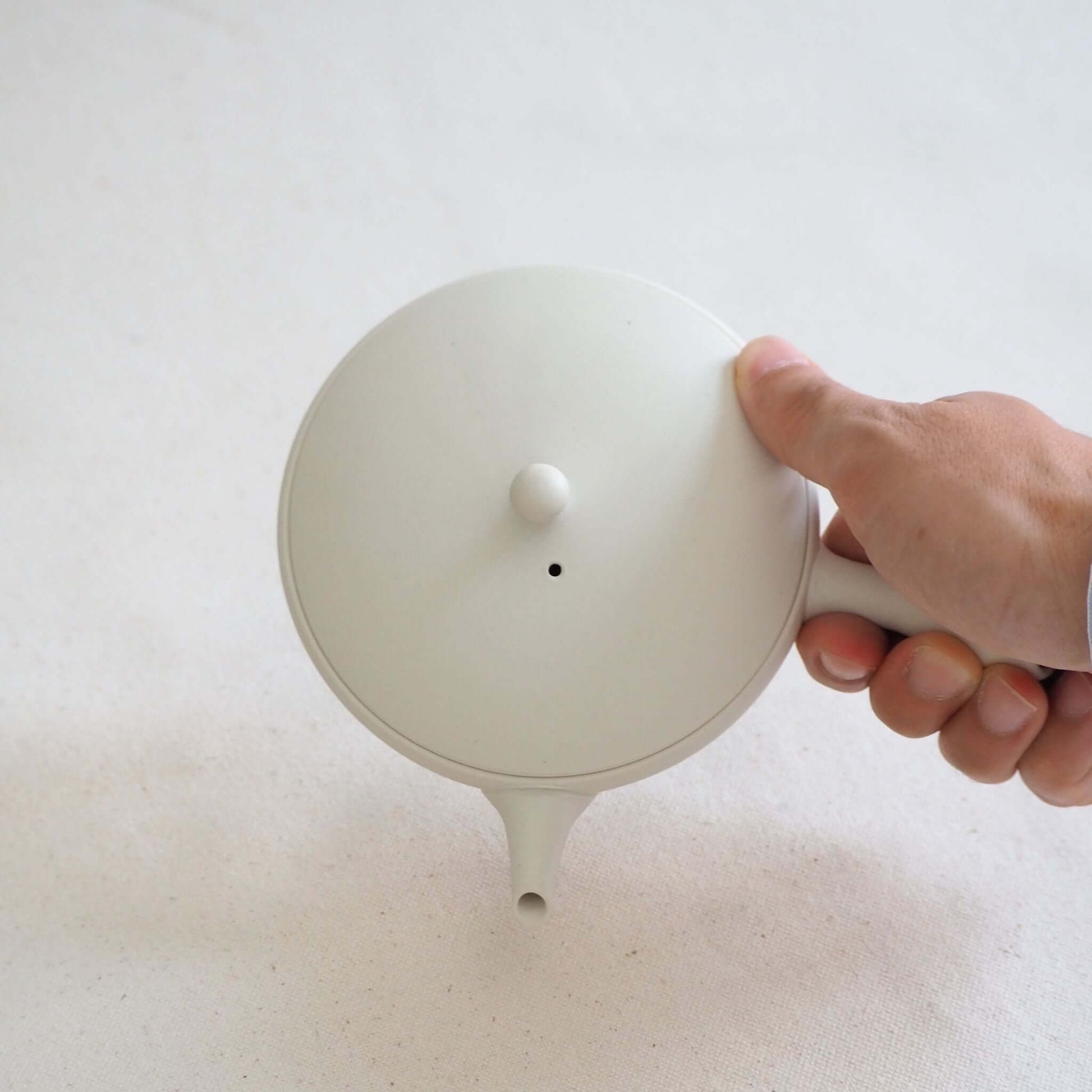 A Tokoname ware white flat teapot is held in the hand and tilted—its soft white appearance is a result of being shaped on a potter's wheel.